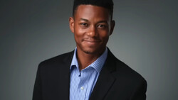Professional headshot of a young man in a black blazer and light blue striped shirt against a neutral gray background.