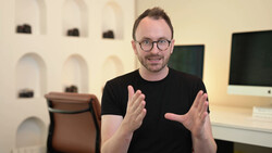 Man in black t-shirt and glasses gesturing while speaking in a modern studio setting.