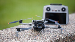 Compact folding drone with camera positioned on gravel surface, remote controller blurred in background against green foliage.