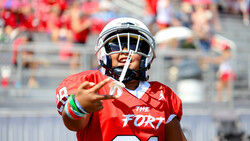 Football player in red jersey pointing toward camera during daytime stadium game.
