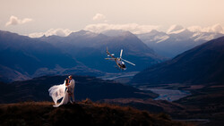 Bride and groom standing on a mountain ridge with a helicopter flying nearby and snow-capped peaks in the distance.