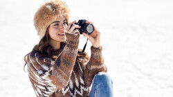 Woman in knit sweater and fur hat holding vintage camera to her face while sitting outdoors.