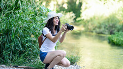 Woman crouching by a riverside holding a camera, wearing a white cap and casual clothing.