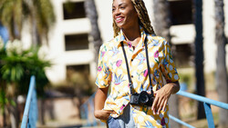 Smiling woman wearing a colorful floral shirt and camera on a lanyard, standing outdoors near palm trees.