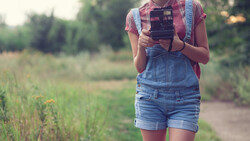 Person in denim overalls and plaid shirt holding a vintage camera in a rural field.