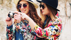 Two women in colorful floral blazers and hats holding a vintage camera between them.