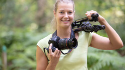 Woman holding professional video camera and binoculars outdoors in natural setting.