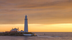 Lighthouse on rocky promontory at golden hour with keeper's house and outbuildings nearby.