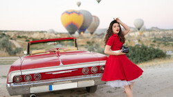 Woman in red dress posing beside vintage red convertible with hot air balloons rising in desert landscape.