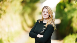 Woman with blonde wavy hair wearing a black cardigan with decorative clasps, standing with crossed arms in an outdoor garden setting.