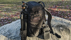 Black camera backpack with tripod attachment sitting on a rock in an outdoor field setting.