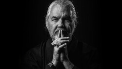 Bearded older man in dark clothing with hands clasped near face against black background.