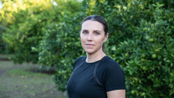 Woman in black t-shirt standing in front of green foliage, looking toward camera.