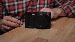 A black Nikon Z mirrorless camera body displayed on a wooden surface, held by a hand wearing a red plaid shirt.