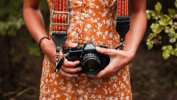 Photographer holding a vintage film camera with decorative shoulder strap, wearing an orange floral dress.