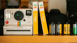 Polaroid One Step+ camera with rainbow stripe branding displayed on wooden shelf alongside Hype film boxes and various film cartridges.