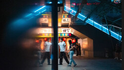 Night street scene with neon signage and blurred figures walking past storefronts.