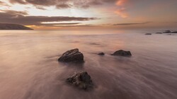 Rocky outcrops emerge from smooth water during golden-hour sunset with dramatic clouds overhead.