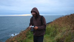 Photographer operating a drone remote controller on a coastal clifftop at dusk.