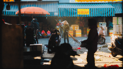 Street market scene viewed through silhouetted foreground figures, with vendors and blue corrugated metal structures under warm daylight.