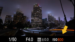 Urban skyline at dusk with illuminated skyscrapers reflected in still water, with camera settings displayed as an overlay.