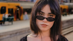 Woman wearing dark oval sunglasses and dark clothing at a train station platform.
