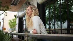 Woman with blonde wavy hair wearing a cream-colored cardigan, seated at an outdoor table with modern architecture in the background.
