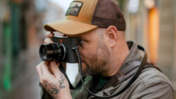 Photographer holding a rangefinder camera up to his eye while wearing a tan and brown trucker cap.