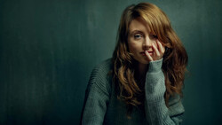 Woman with long reddish-brown hair wearing a gray-green sweater, resting her face against her hand against a teal background.