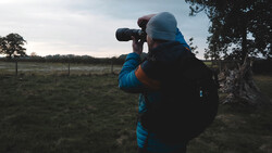 Photographer in blue jacket and beanie using telephoto lens in rural pasture at dusk.