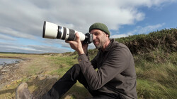 Photographer holding a telephoto lens to his eye while crouching on a hillside near water.