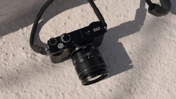 Black rangefinder camera with attached lens on white textured surface, casting sharp shadows in bright sunlight.