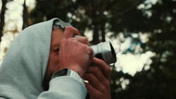 Photographer holding binoculars to eyes while wearing a hooded sweatshirt outdoors.