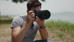 Male photographer holding a DSLR camera with telephoto lens, composing a shot outdoors.