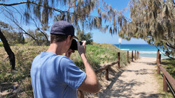 Photographer with camera raised to face, composing a shot down a sandy beach path lined with wooden railings and coastal vegetation.
