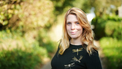 Woman with reddish-blonde wavy hair wearing a black embroidered top, photographed outdoors with blurred green foliage background.