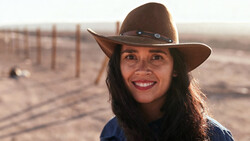 Woman wearing a brown cowboy hat and blue shirt, smiling at the camera in a desert landscape at golden hour.