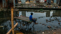 Child in blue shirt sitting on colorful cart in shallow water amid urban debris and waste.