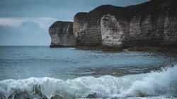 Dramatic limestone sea cliffs rising from foaming ocean waves under a partly cloudy sky.