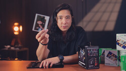 Creator holding instant film photograph at desk surrounded by camera equipment and lighting.