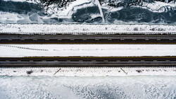Aerial view of snow-covered landscape with railroad tracks and forested areas separated by white fields.