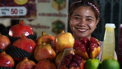 Smiling vendor at a fruit market stand displaying pomegranates and other fresh produce.