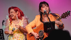 Two performers on stage with microphones; one in a yellow polka dot dress with red hair, the other in an orange jacket playing an acoustic guitar.