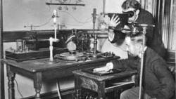 Photographer using a large format camera mounted on a tripod to photograph a subject seated at a table below.