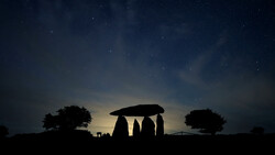 Silhouette of Stonehenge monument against a twilight sky with scattered stars and trees on the horizon.