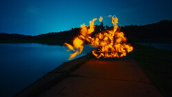 Bright bonfire burning on a beach at dusk, reflected in calm water with dark hills silhouetted against blue twilight sky.