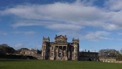 Stone castle with crenellated towers set on manicured grounds under partly cloudy sky.