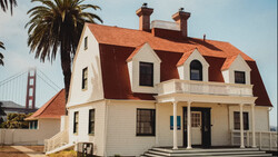 White Victorian house with red mansard roof and palm tree beside Golden Gate Bridge in background.