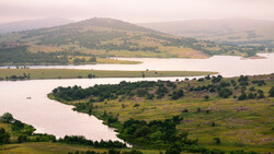 Meandering river through rolling green valley with conical mountain in misty distance.