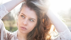 Woman with long brown hair backlit by golden sunlight, holding a white lace fabric above her head.